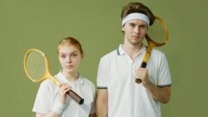 A young man and woman in sportswear pose with vintage squash rackets in a studio setting.