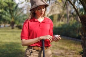 A woman golfer holds a golf ball on a course, dressed in a red polo and brown hat on a sunny day.