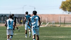 Teen boys playing soccer on a green field under a clear blue sky, wearing team jerseys.