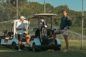 Two men at a golf course with carts and equipment. Sunny outdoor setting.