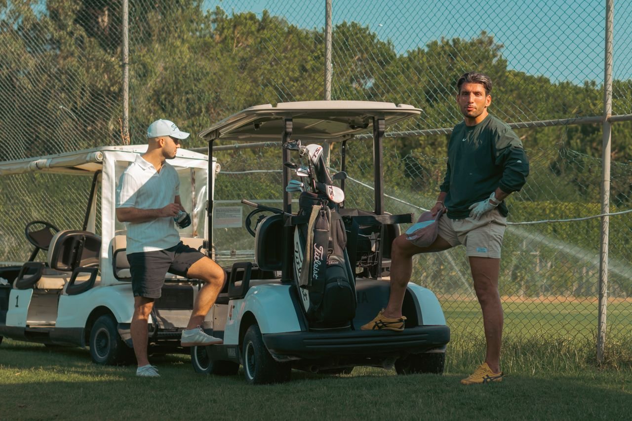 Two men at a golf course with carts and equipment. Sunny outdoor setting.