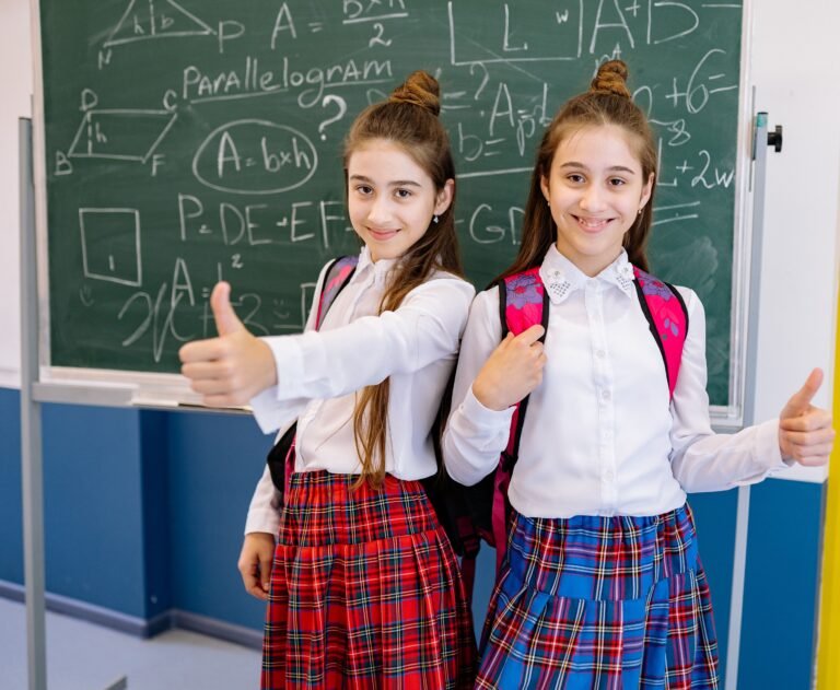 Two cheerful schoolgirls in uniforms giving thumbs up in front of a chalkboard filled with math equations.