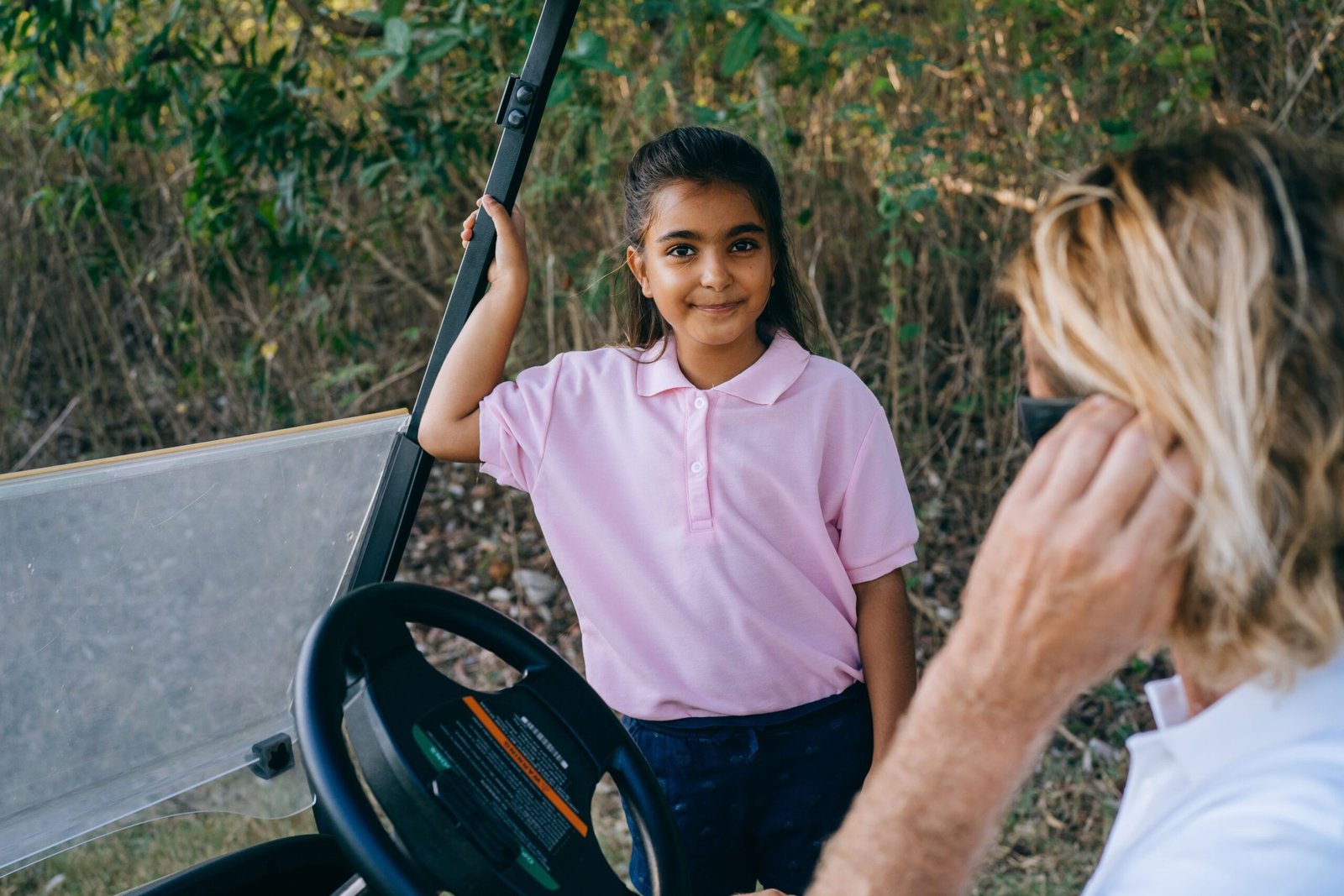 Young girl smiling beside a golf cart in a sunny outdoor setting with trees.